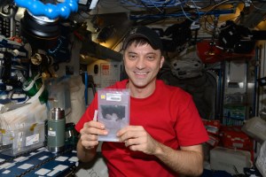 Dominick, wearing a red short-sleeved t-shirt and a black ball cap, smiles at the camera and holds up in both his hands a sandwich-sized plastic zipper bag containing a black powder.