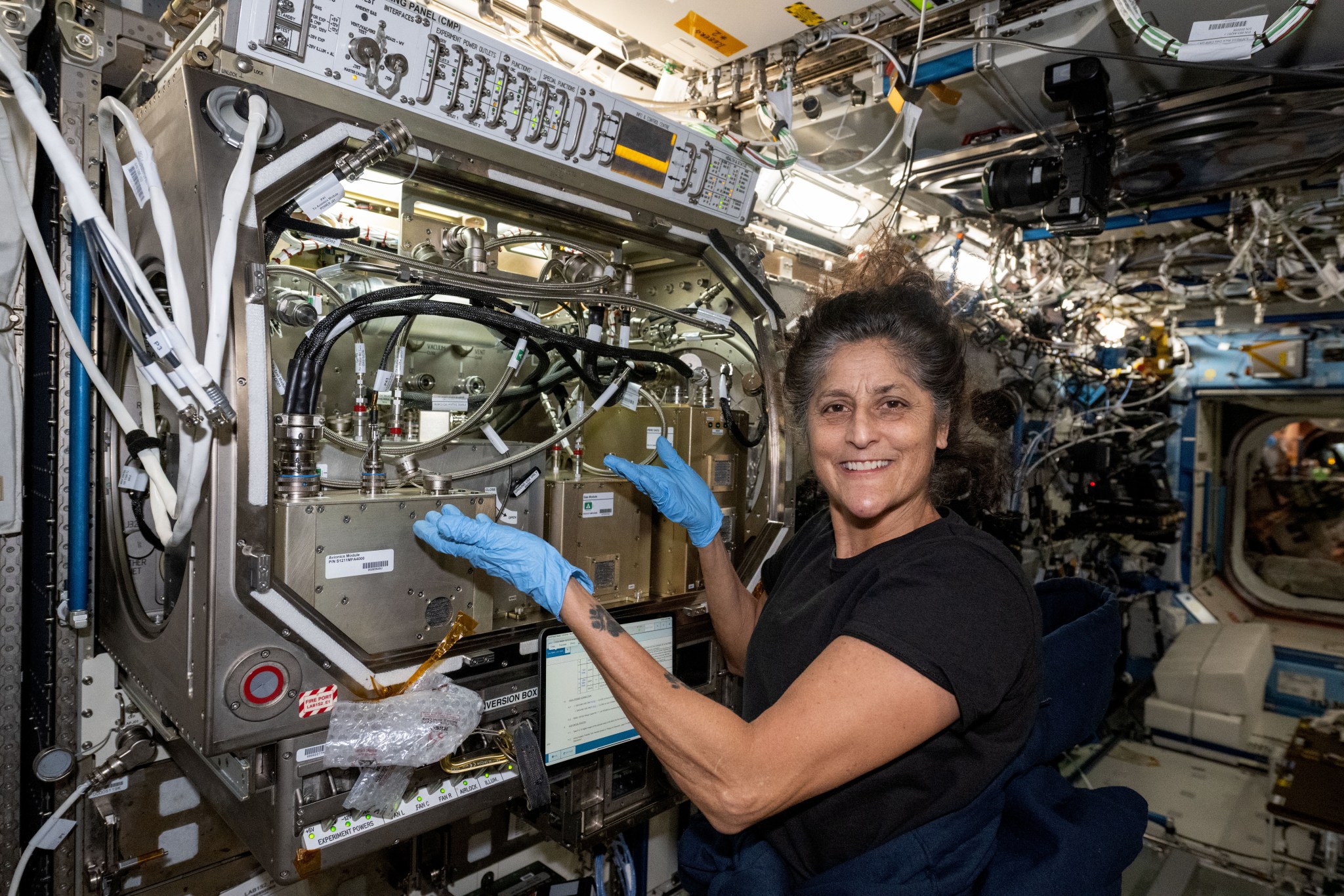 Williams, vestida con una camiseta negra y guantes de látex azules, sonríe a la cámara y señala hacia el hardware para el experimento. A su izquierda, al fondo, hay varias cajas de metal con diferentes mangueras y cables negros y plateados que unen las cajas a los equipamientos. Hay un panel de control blanco en la parte superior con diferentes etiquetas e interruptores.