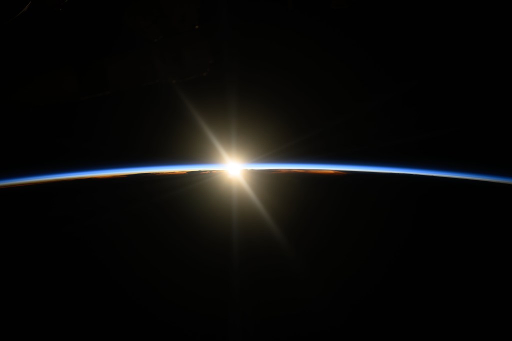 The beginning rays of an orbital sunrise illuminate Earth's atmosphere, a slight, blue arc across the center of the image. Earth is below, totally dark, matching the blackness of space above.