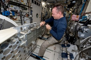 NASA Astronaut Butch Wilmore works on a large silver box in front of him using a screwdriver. Wilmore has his feet under two blue bars on the floor to prevent him from floating aware. He is earing khaki pants and a navy long-sleeve shirt.