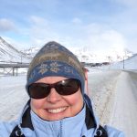 A selfie of a woman in a blue coat, knitted beanie, and sunglasses standing in front of an icy background. She appears to be standing on the side of a slushy, cleared road and snow-covered mountains and cloudy blue skies are behind her.