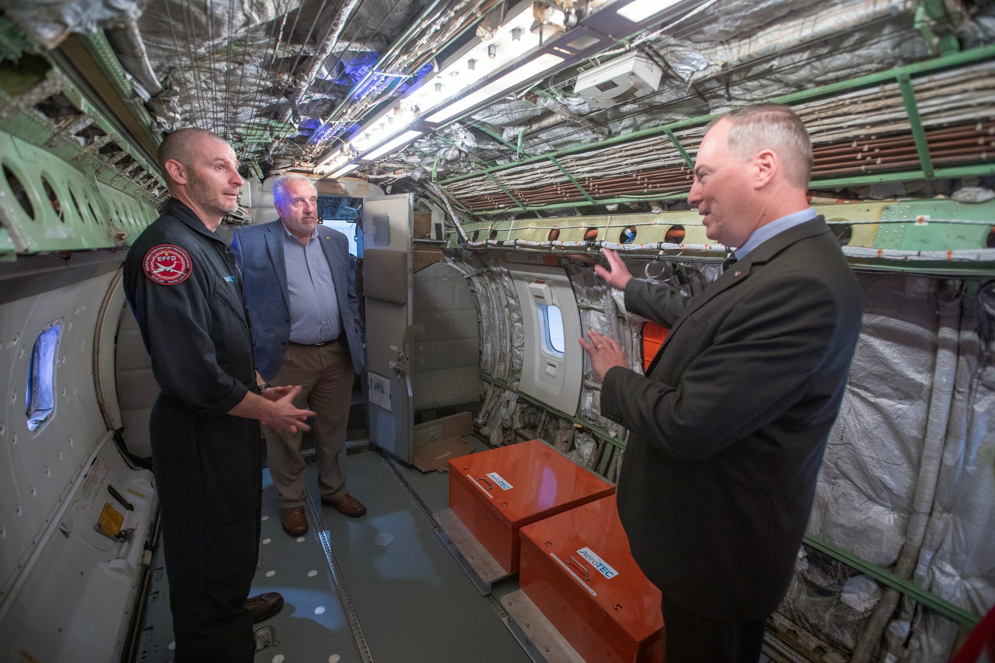 Three men engage in a conversation inside a hollow cabin surrounded by various hardware, insulation, lights, and red boxes along the floor used for storing the aircraft’s battery packs.