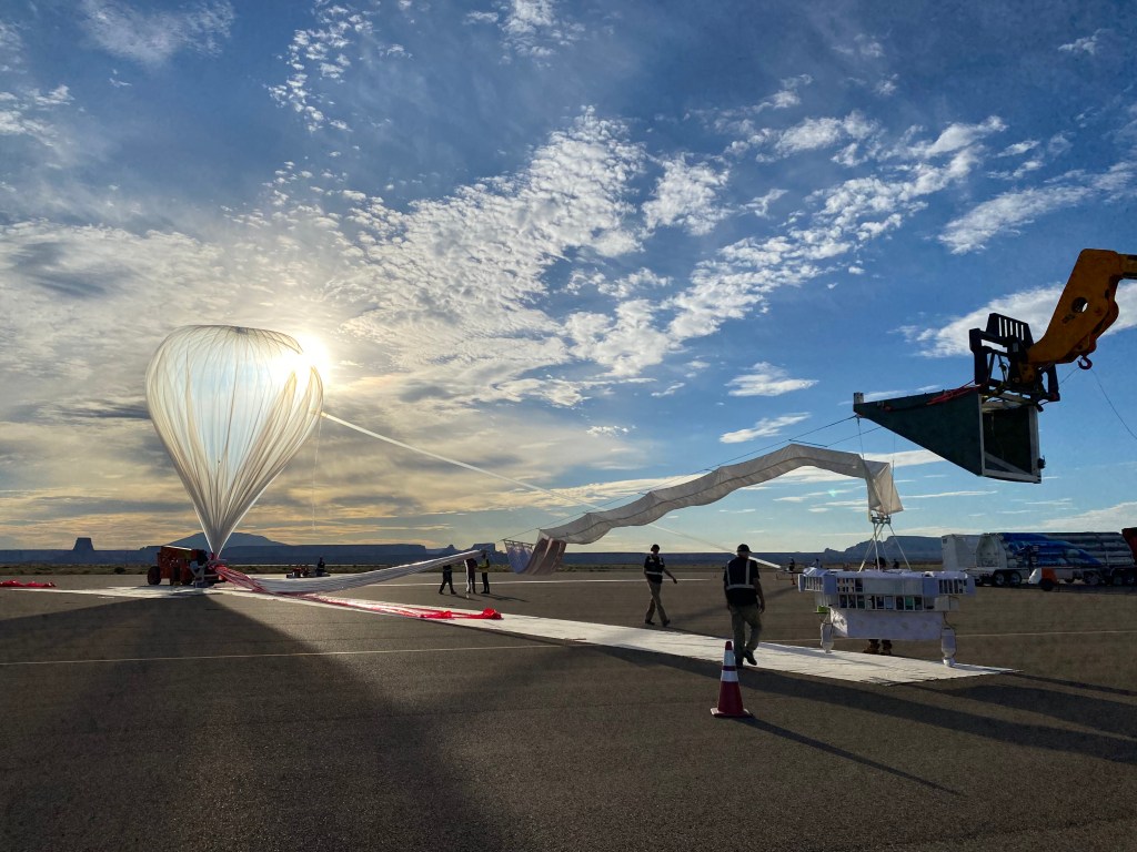 An inflated high-altitude balloon with the sun shining behind it sits on the tarmac with a long tether leading to its payload.