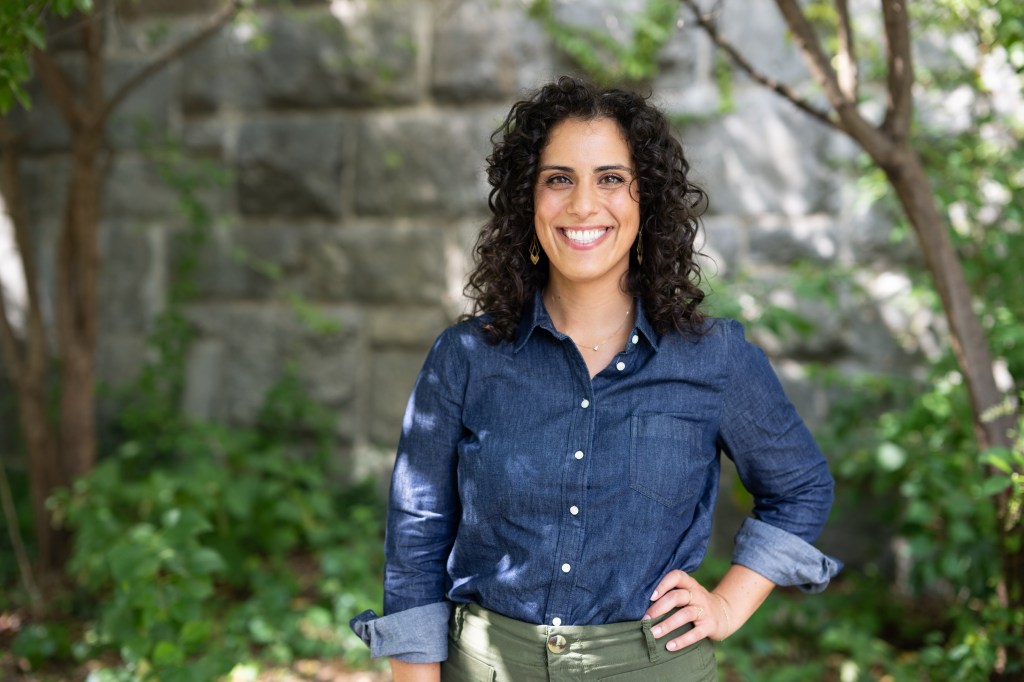 Emily Furfaro, a woman with shoulder-length dark brown hair and brown eyes smiles brightly at the camera while wearing a blue jean button up shirt and olive pants.