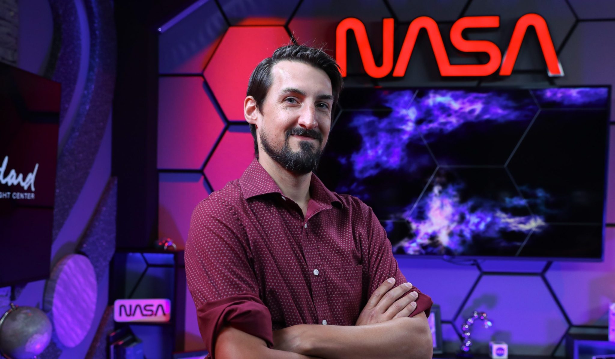Rob Garner stands in the Goddard TV studio next to a galaxy image and a red NASA worm sign. The wall is made up of hexagons and lit by the sign and a purple light. Garner is smiling and is wearing a red button down shirt with white dots and stands with his arms folded across his chest. 