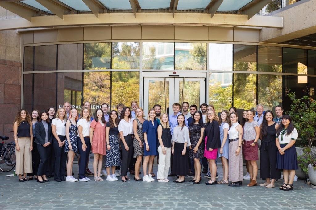 A group of 24 college students, eight mentors, and additional staff pose together in front of a glass building. All participants are wearing professional clothing in a variety of colors.