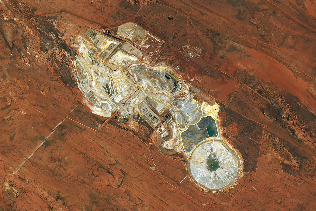 An overhead view of a mine in the dry outback of Australia. Most of the image is dominated by rusty red soil. Miners have excavated the soil in places, revealing an off-white color in these open pits. Some water is visible as well, in ponds.