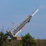 A sounding rocket is being raised on the launch rail, it is still in a horizontal position. The red a silver rocket contrasts against the bright blue sky behind, and green foliage can be seen at the bottom of the photo
