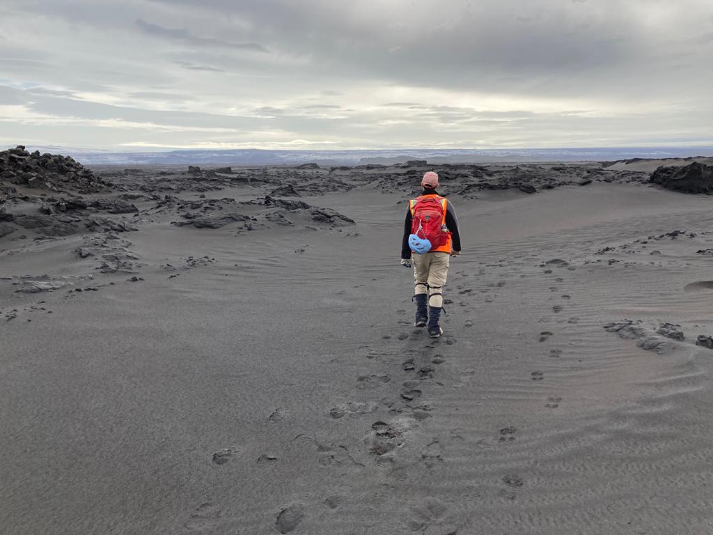 Zachary Morse walks on soft grey-brown ground where his footprints are visible. He wears boots, tan pants, a safety vest, and a red backpack. There is a blue helmet clipped to his backpack. The photo is taken from behind.