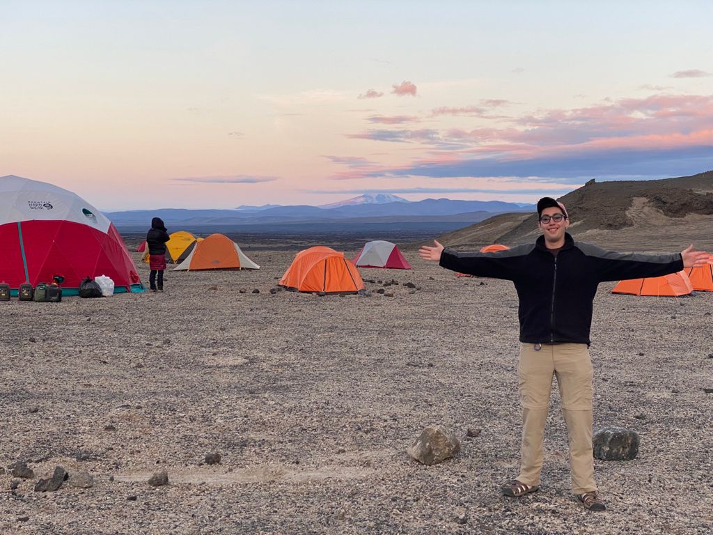 Zachary Morse stands with his arms outstretched wearing a hat, jacket, and tan pants in front of several red and orange tents with a mountain in the background. The sky is pink and blue.  