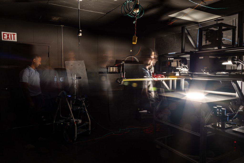 A long-exposure photo of a dark room painted all black, with a man in a light blue shirt moving from left to right. On the right is a bright light coming out of a table.