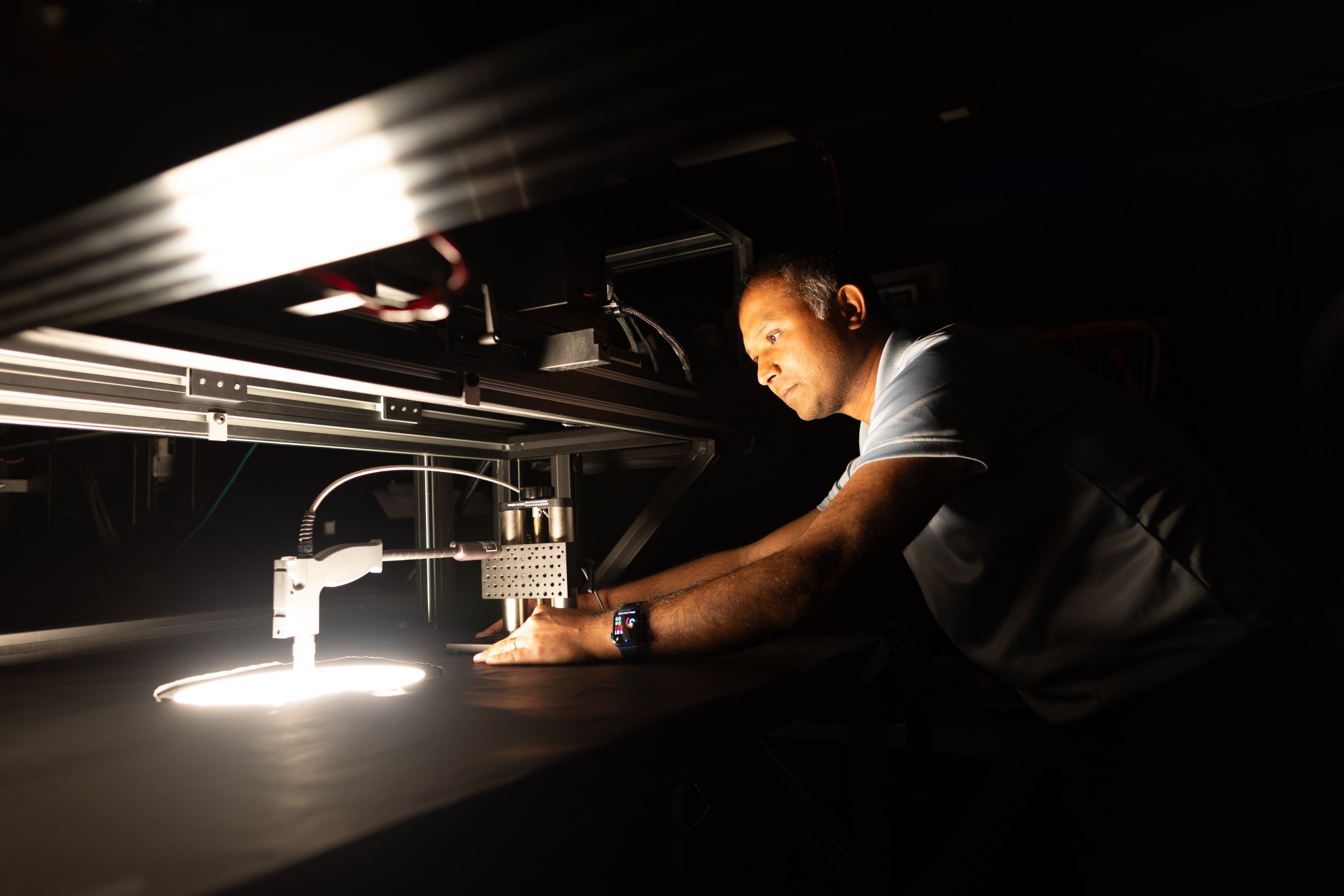 A middle-aged indian man in a blue short-sleeved shirt leans over a table with a circular hole cut in the middle. The room is entirely dark except for a bright light coming out from the hole, illuminating the man's face.