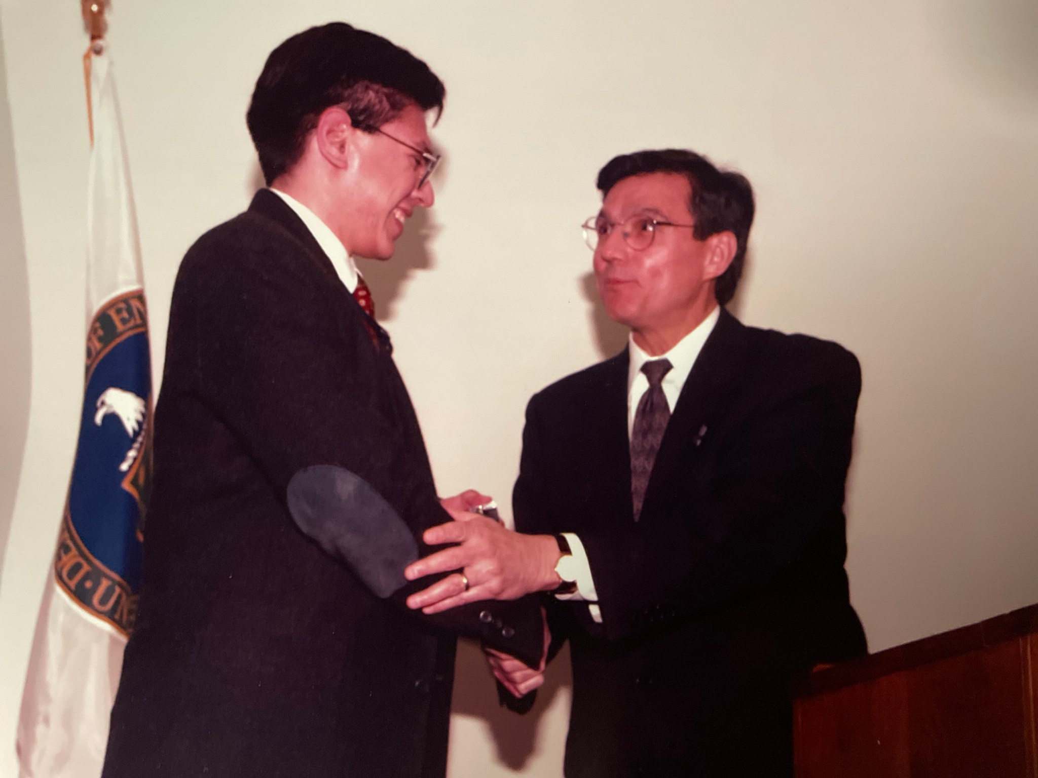 Two men in suits exchange a handshake during a formal event. One man, smiling, holds an object wrapped in a dark cloth, while the other looks on with a slight smile. Behind them is a flag with a seal.