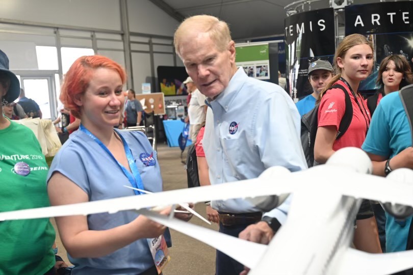 NASA employee, left, talks with the NASA administrator, right, while standing behind a white plane model. People are walking around in the background.