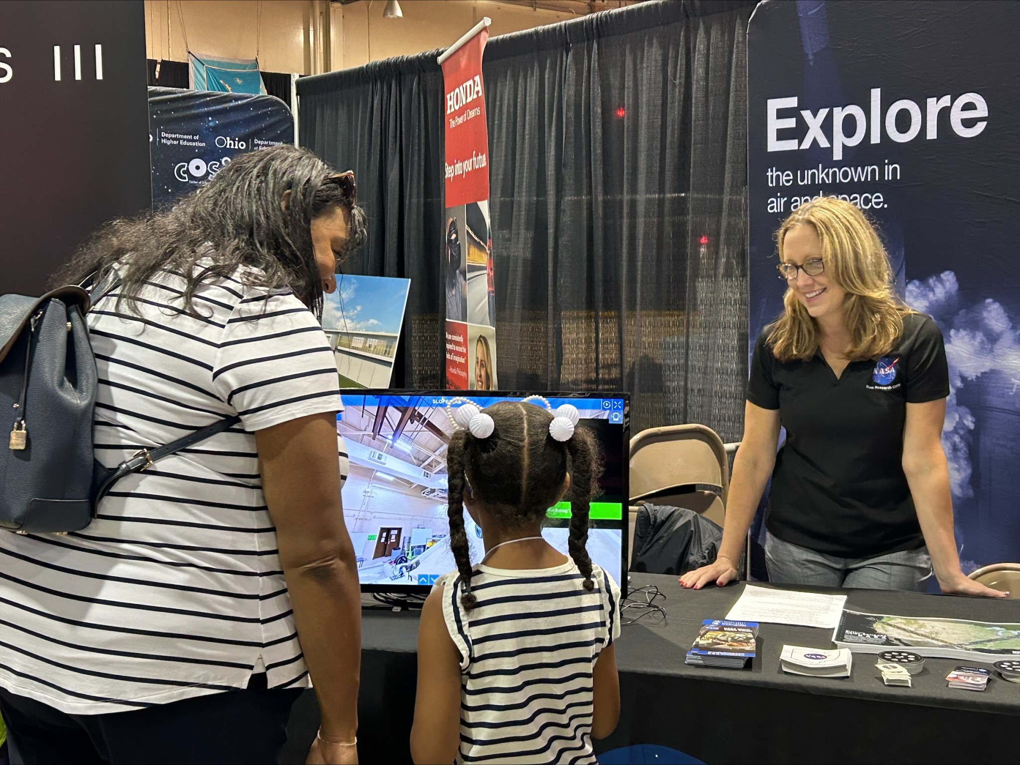 A mother and daughter look at a monitor that shows a virtual tour of a facility at NASA’s Glenn Research Center. A Glenn specialist stands behind a table and explains details about the video.