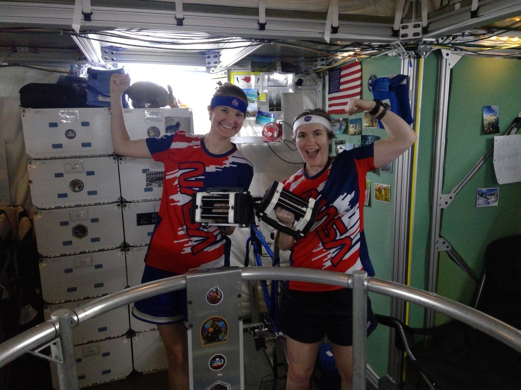 Two individuals wear red, white, and blue athletic shirts and headbands while raising their fists in excitement and holding exercise equipment inside HERA.