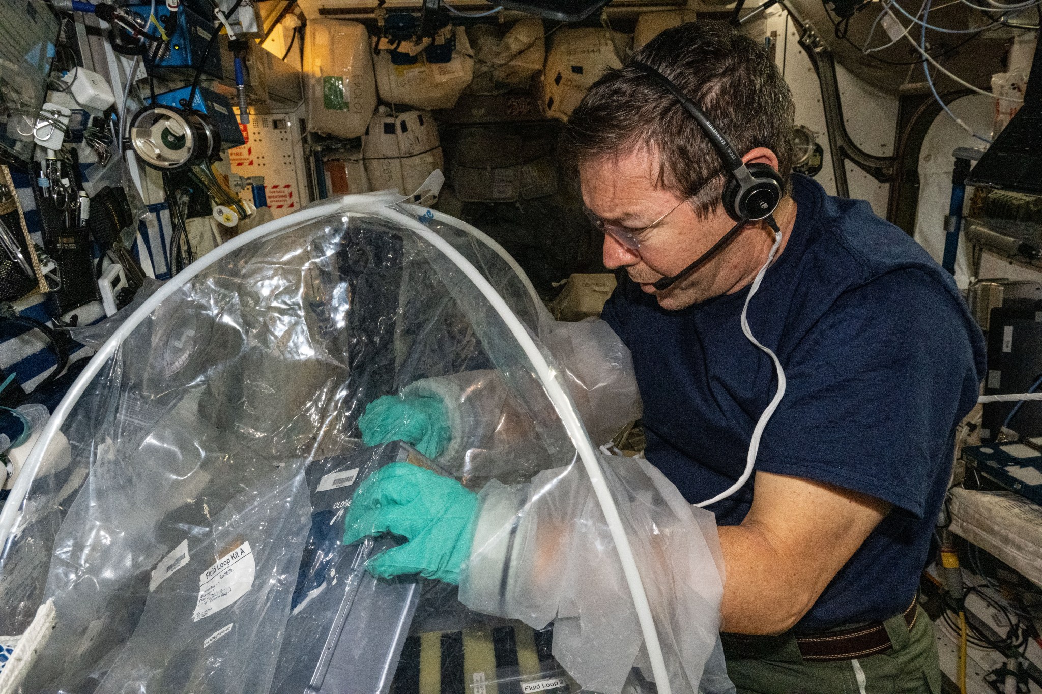 Barratt is wearing a dark blue t-shirt, green pants, and a headset. He is peering at his hands inside the sleeves of a portable glovebox, holding a sample cassette, a silver box the size and shape of a briefcase.