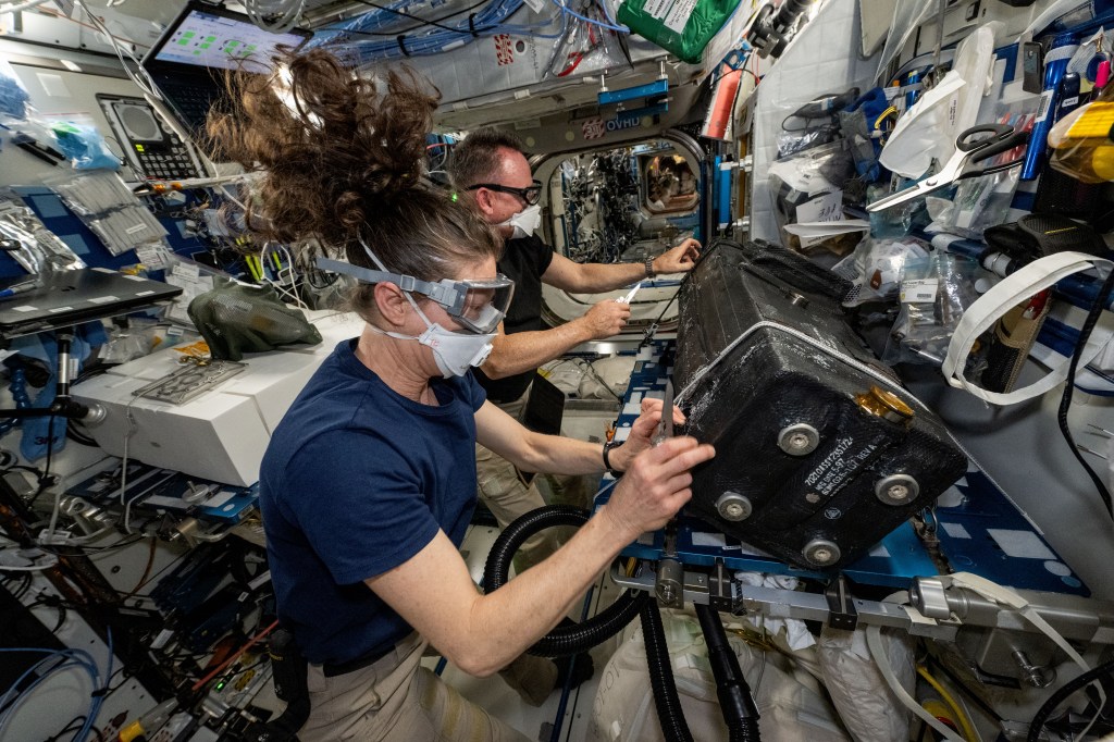 NASA astronauts Tracy C. Dyson (foreground) and Butch Wilmore wear personal protective equipment and clean the inside of the carbon dioxide removal assembly that is part of the International Space Station’s Air Revitalization System located inside the Tranquility module.