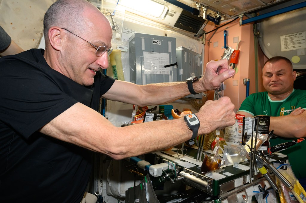 Expedition 71 Flight Engineer (from left) Don Pettit from NASA demonstrates filling a specially-made coffee cup from a drink bag to Expedition 71 Flight Engineer Alexey Ovchinin from Roscosmos. The coffee stays inside the cup due to surface tension, a property that enables liquids to resist an external force, such as microgravity, and adhere, or stick to a surface.