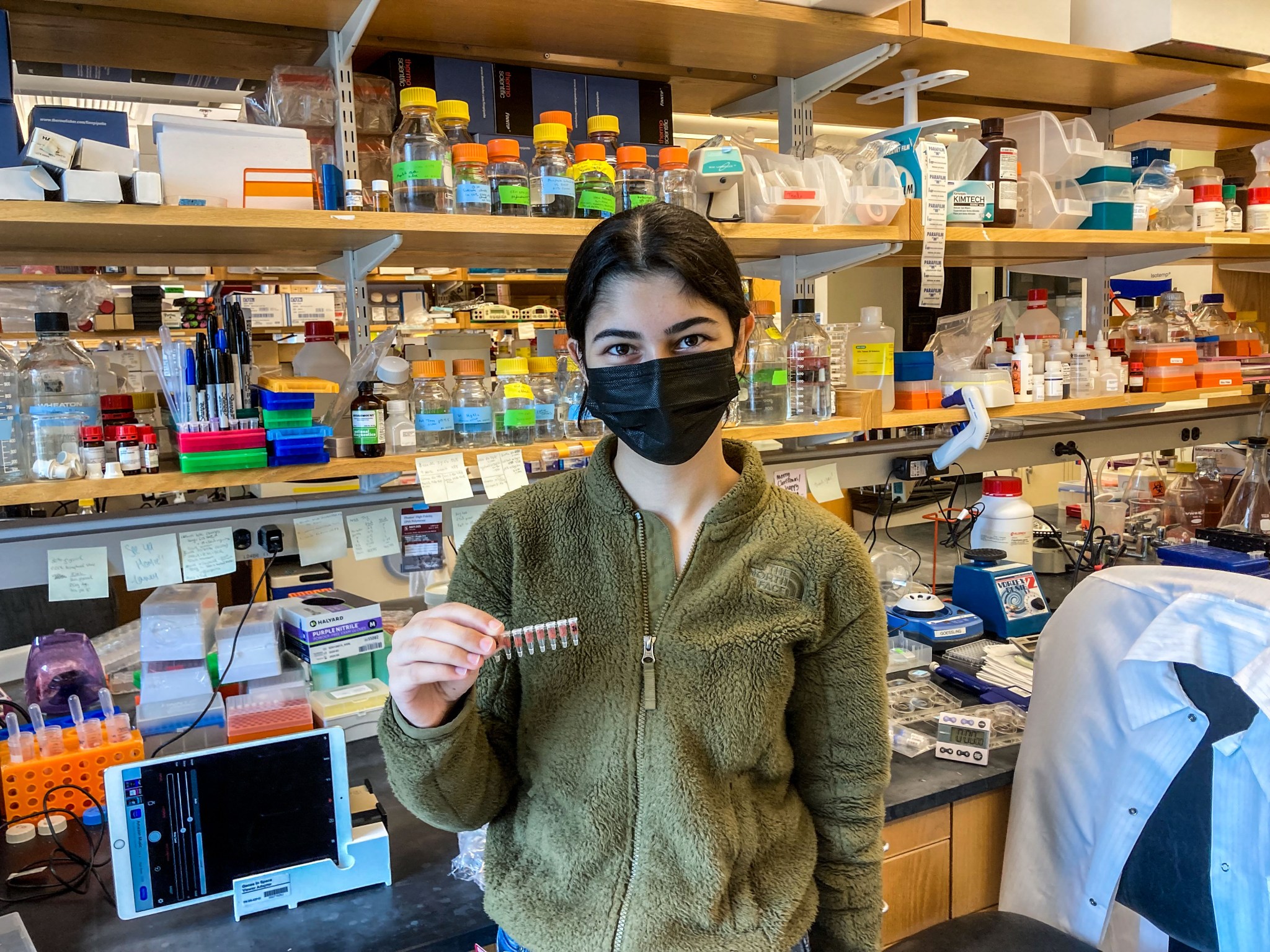 Kocalar is wearing a green fleece jacket and a black face mask, with her hair pulled back in a ponytail. She is facing the camera and holding a sleeve of clear plastic sample tubes. To her right is a device with a view screen and to her left a chair with a white lab coat draped over it. Behind her is a lab counter and shelves filled with a variety of laboratory equipment.