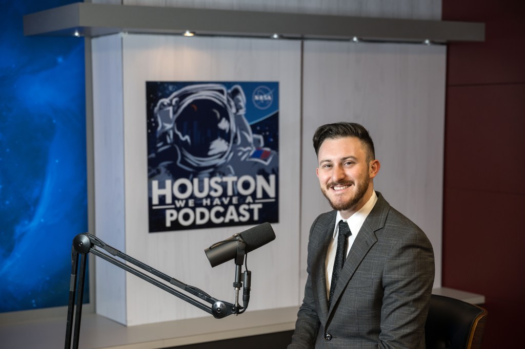 Public Affairs Manager Gary Jordan smiles at the camera from where he sits in Studio B at NASA's Johnson Space Center. He sits in front of a microphone. A poster in the background says "Houston We Have a Podcast."