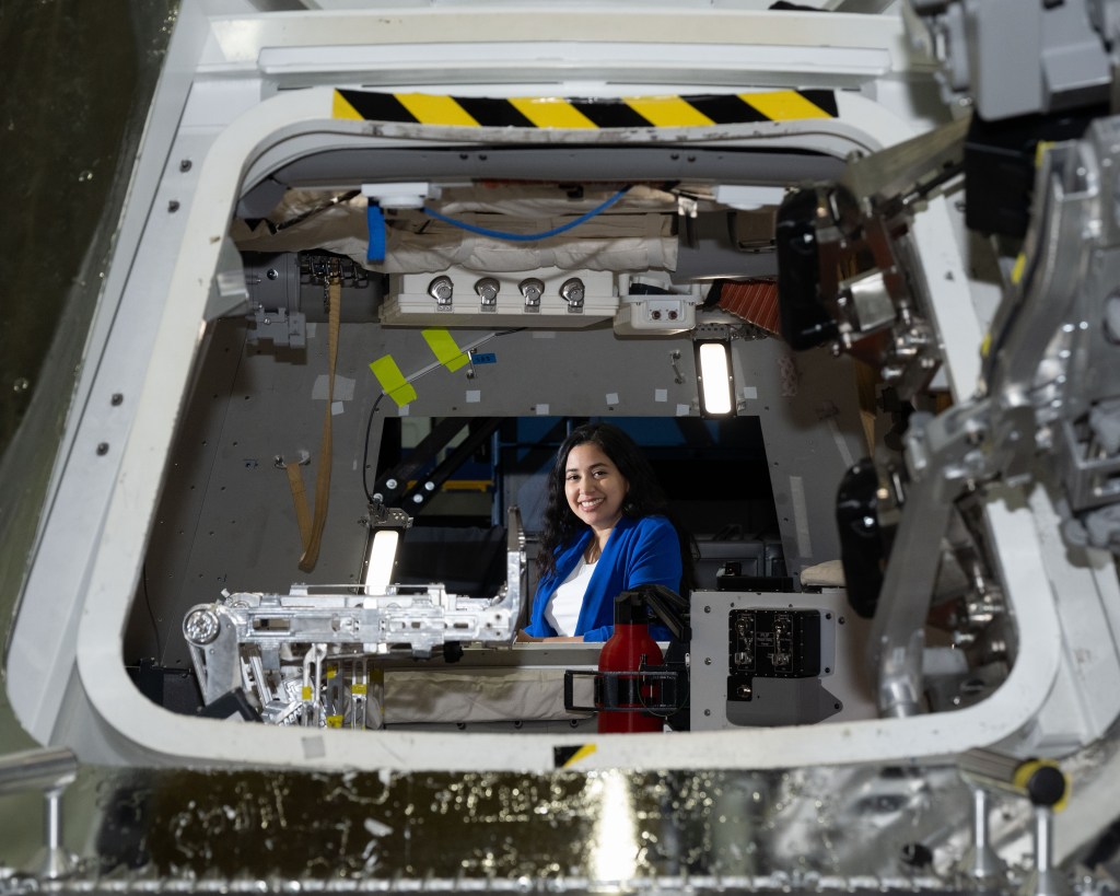 Zaida Hernandez, a hispanic woman with long wavy brown hair and dark brown eyes, smiles brightly at the camera while sitting inside a mock up of NASA's Orion Crew Capsule. She's wearing a blue blazer and flight hardware is seen all around her.
