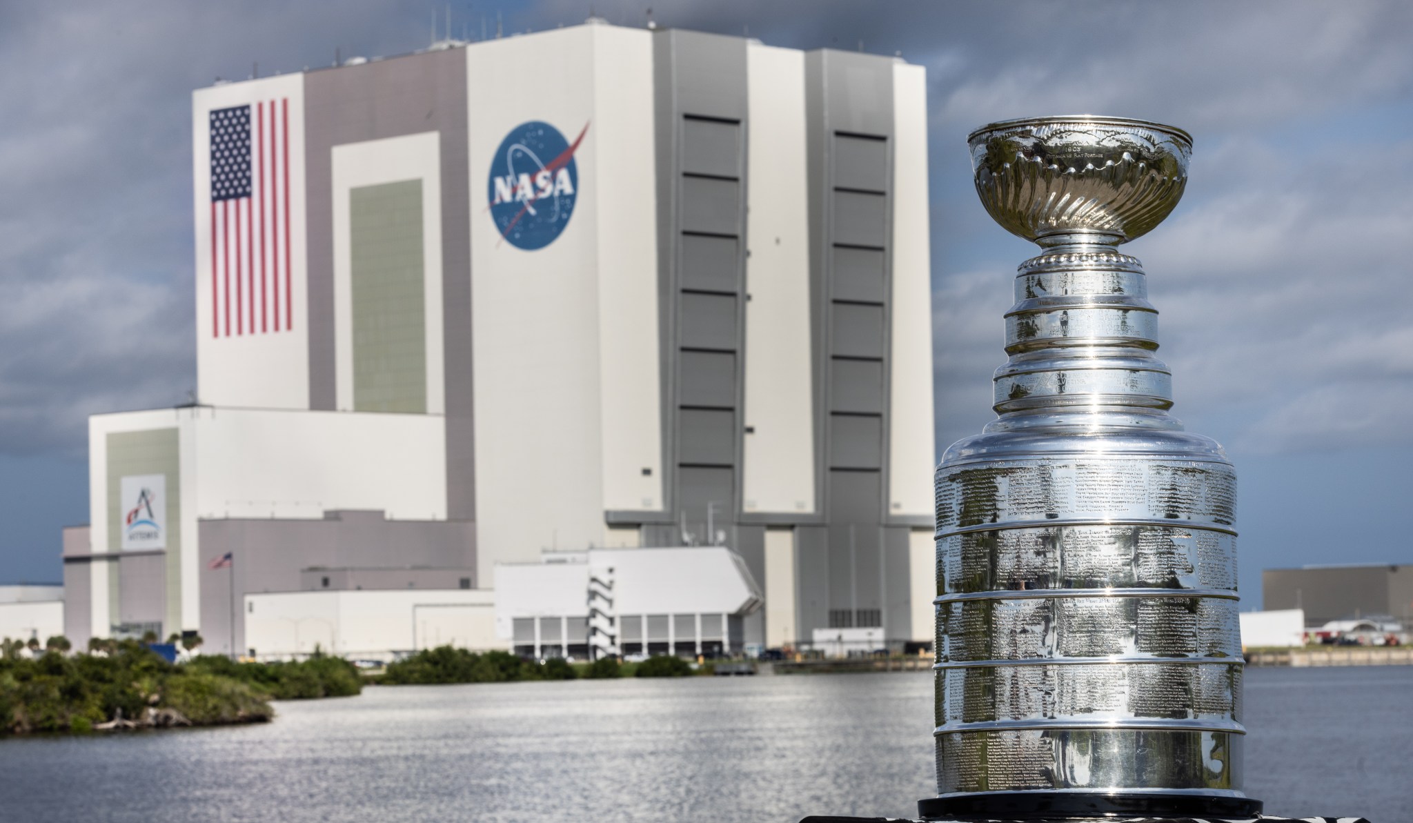 The silver Stanley Cup is in the foreground at right. The Stanley Cup looks like a large silver bowl set on top of a stacked column of thick silver layers. There are words engraved on the Cup. Behind it is a body of water and the Vehicle Assembly Building at NASA's Kennedy Space Center, a massive, boxy gray and white building.