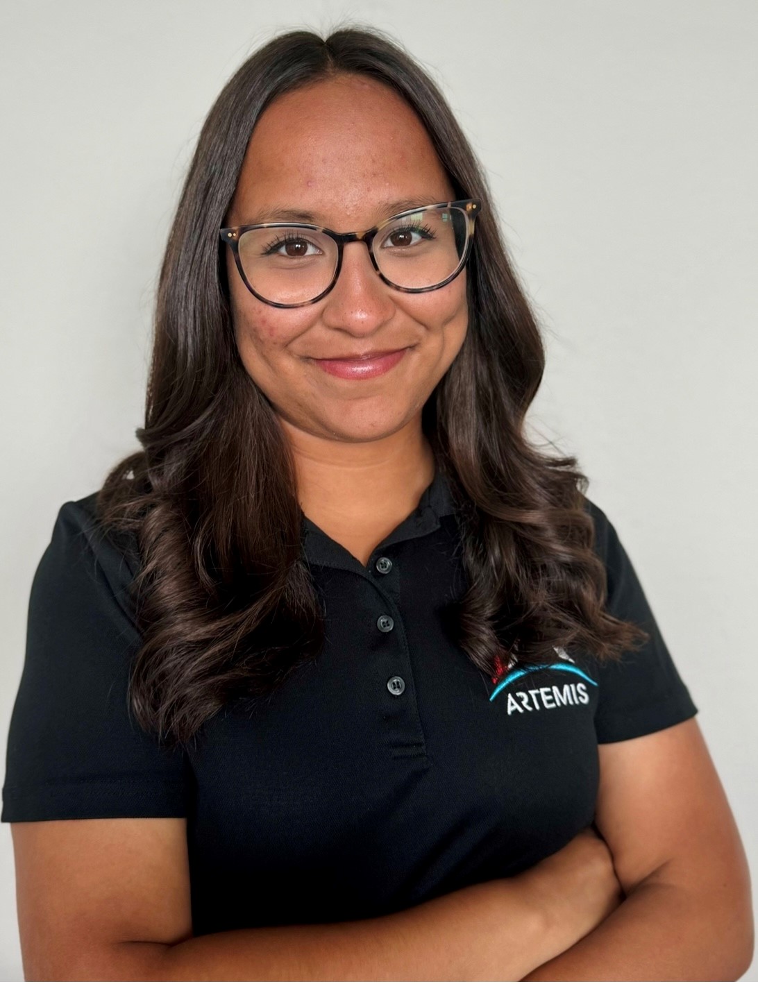 A woman with long, dark brown hair, wearing black-framed glasses and a black Artemis polo shirt, smiles while standing with her arms crossed.