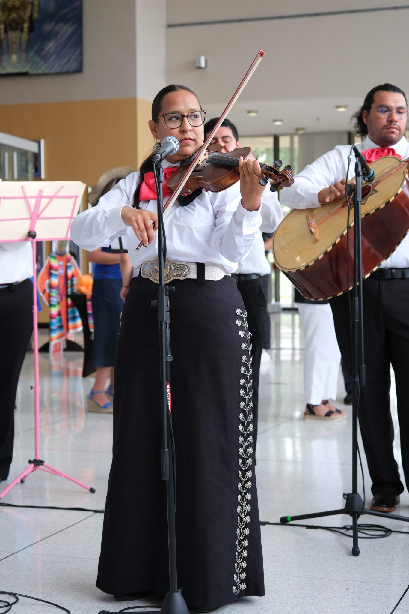 A female musician dressed in traditional mariachi attire, consisting of a white shirt, black skirt with silver embellishments, and a red necktie, plays the violin while singing into a microphone. She stands in front of a band with other mariachi members, including a drummer in the background, during a performance in an indoor venue.