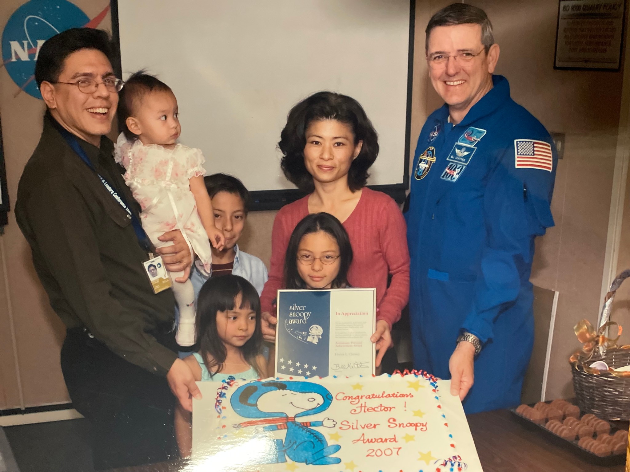 A family poses together with an astronaut in a NASA jumpsuit during a Silver Snoopy Award ceremony. The father holds a small child, while the mother displays the Silver Snoopy Award certificate. A large decorated cake featuring Snoopy and the text ‘Congratulations Hector! Silver Snoopy Award 2007’ is placed in front of them.