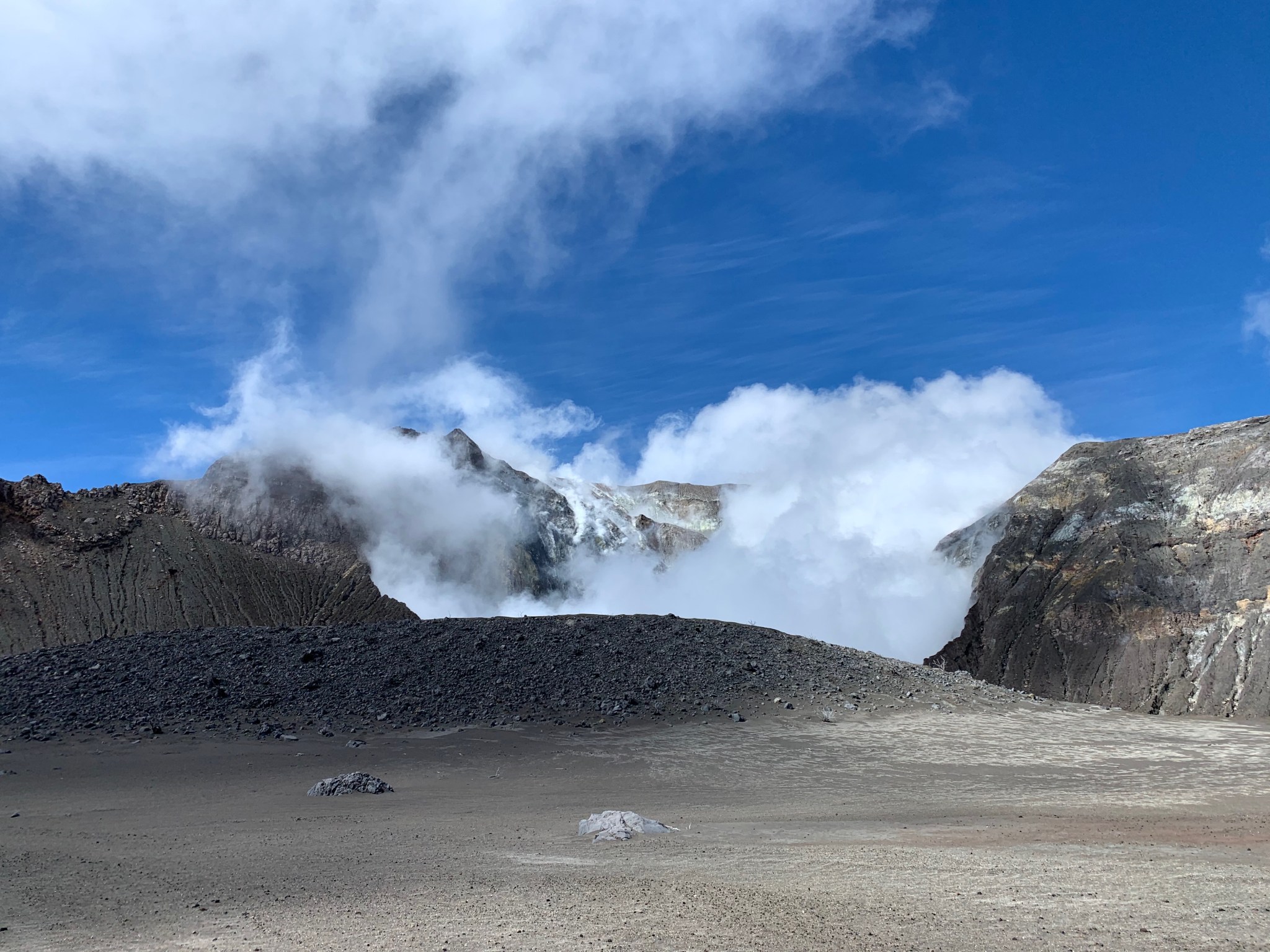 A grey mountain rises in the middle of the photo into a blue sky, with bits of white clouds swirling around the peak. In the foreground, the same gray rock makes a flat plane of sand.