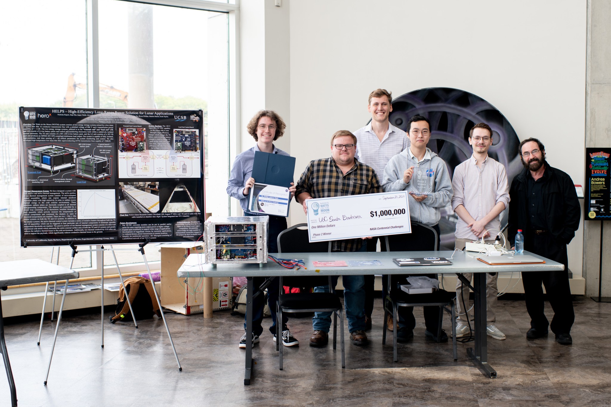 At the Great Lakes Science Center in Cleveland, Ohio, Team H.E.L.P.S. (High Efficiency Long-Range Power Solution) from The University of California, Santa Barbara pose with their $1 million check and the power transmission and energy storage hardware that won them the grand prize in the four-year competition. The team is comprised of six faculty researchers at UC Santa Barbara, led by team lead Dr. Philip Lubin (far right).