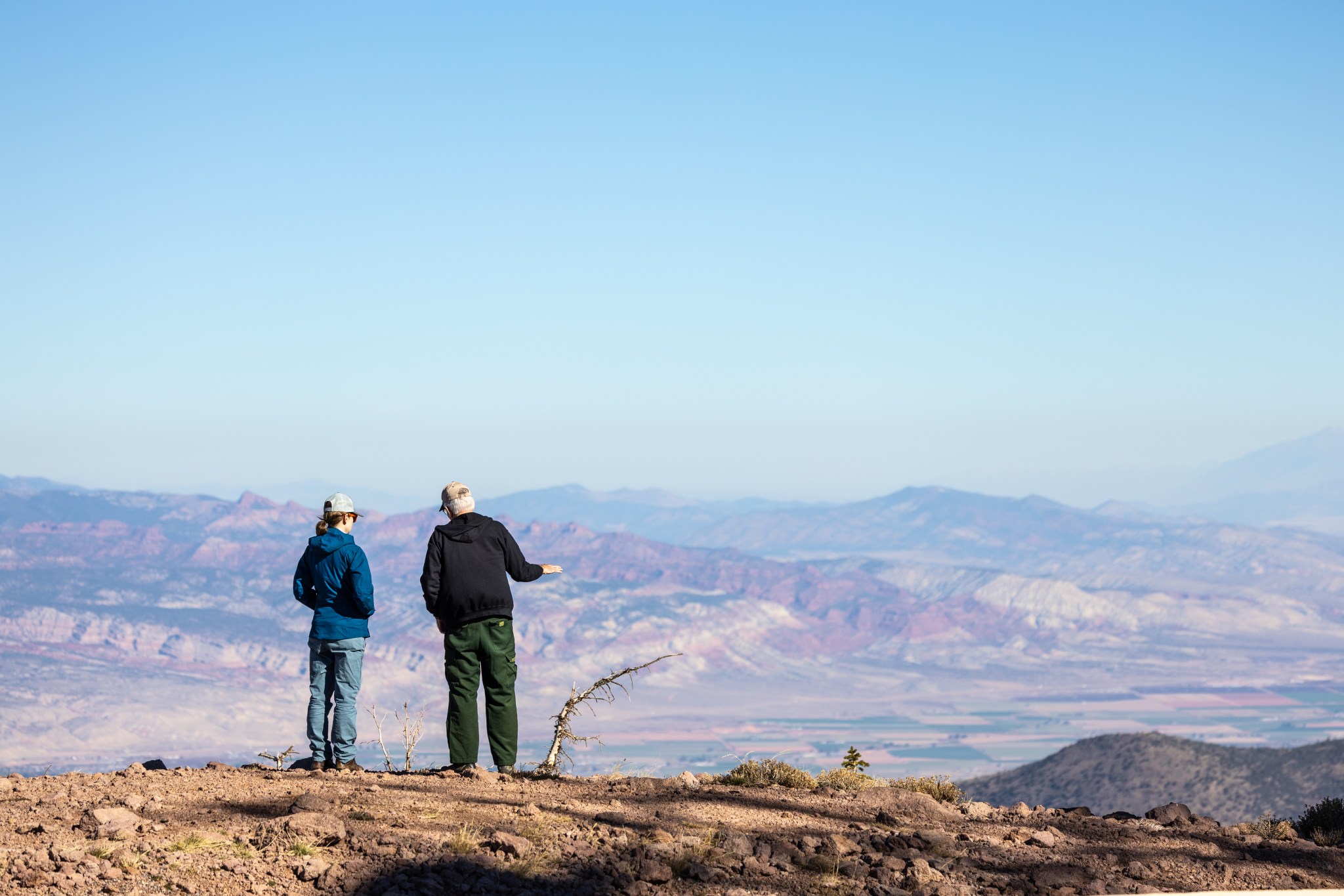 A white woman in a blue jacket and jeans stands next to a white man in a black sweatshirt and green pants, looking out over a valley. They stand on brown dirt, and the sky in front of them is clear and blue, the mountains a darker shade of blue and purple.