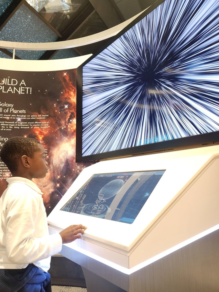 alt="A young student uses an interactive kiosk at the NASA Goddard Visitor Center to build an Exoplanet. A large video screen also shows star streaks as space flight is simulated."