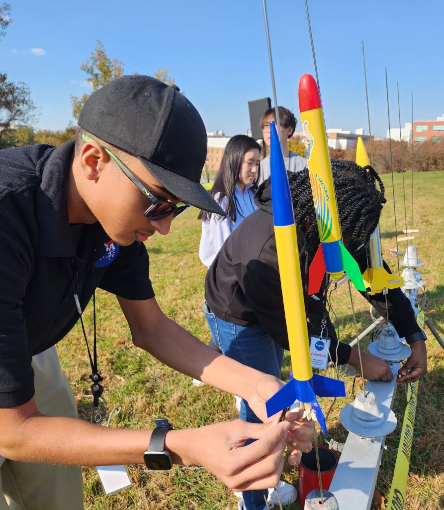 A group of volunteers assist in launching model rockets at the NASA Goddard Visitor Center. Several multicolored rockets are seen on a launching rack on the right of the image, while four high school students assist with the wiring as preparation to launch.