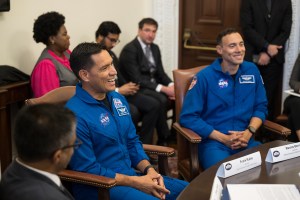 Two Hispanic men in blue jumpsuits with NASA patches on them sit at a table, smiling at people off screen. There are people of different ethnicities in the background.
