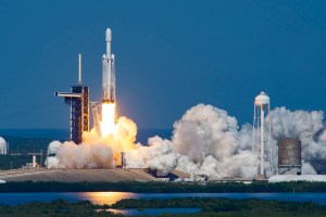 A white rocket lifts off from the launch pad, spewing billowing clouds of white vapor horizontally along the ground. A relatively short column of flames erupts from below the rocket just after liftoff. The early afternoon sky and the water in the foreground are similar, vibrant shades of blue.