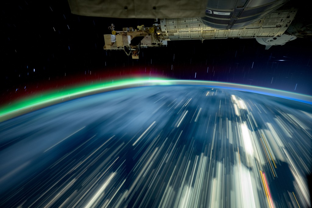The top third shows part of the space station in focus. The bottom two thirds show the Earth with long streaks of white light against the blue of Earth captured using long-exposure photography. To the left is a green and red aurora.