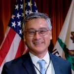 Portrait of a smiling William N. Chan. The American and California flags can be seen in the background.