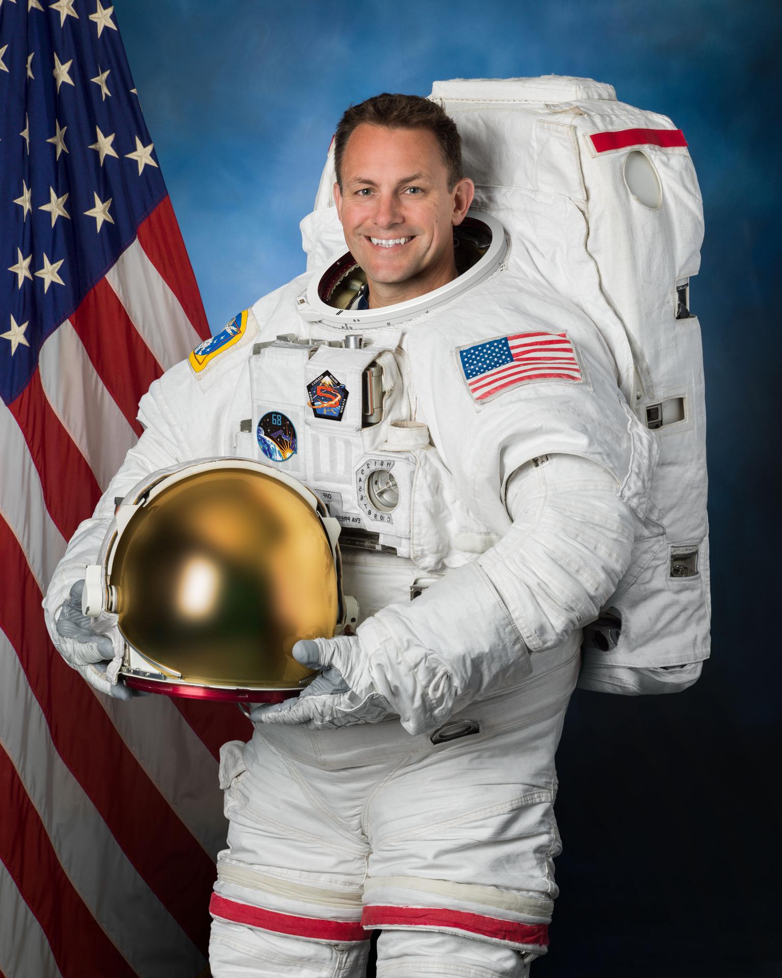 NASA astronaut Josh Cassada poses in front of an American flag in his white extravehicular mobility unit spacesuit and holding his helmet in hands.