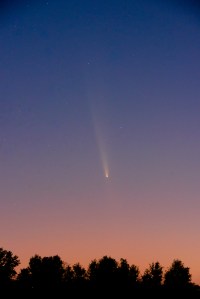 The night sky goes from blue to pink as you look from top to bottom. In the middle is a comet; it looks like a relatively bright yellow dot with a long, diffuse yellow tail. Some stars are visible in the blue portion of the sky.