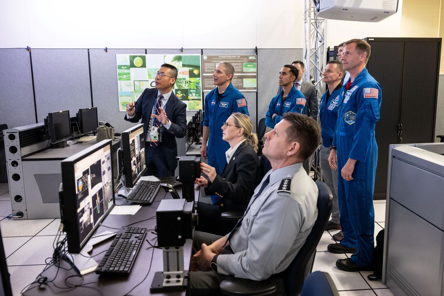 A group of NASA astronauts and officials, including a military officer, observe a presentation on computer screens in a training lab.