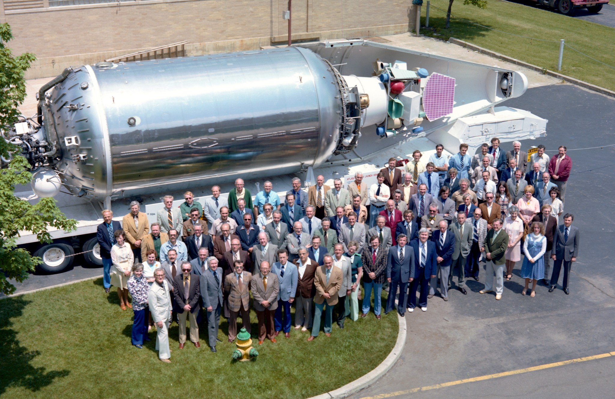 An overhead view of a large group of people wearing professional clothing who stand outside in front of a large, silver rocket model.