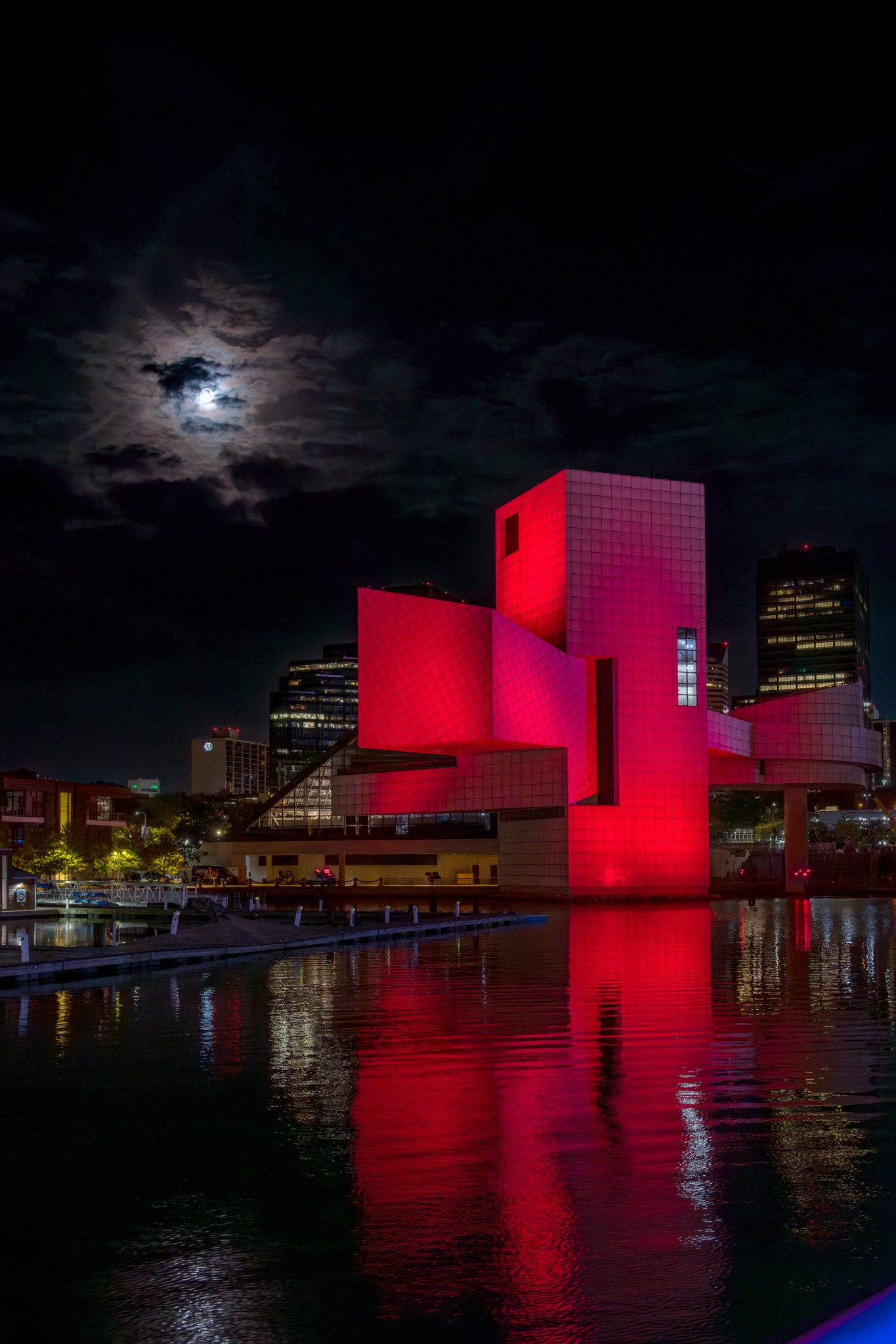 This shot shows the Moon above the colorfully illuminated (in red) Rock & Roll Hall of Fame in downtown Cleveland. The image is mirrored in the water next to the building.