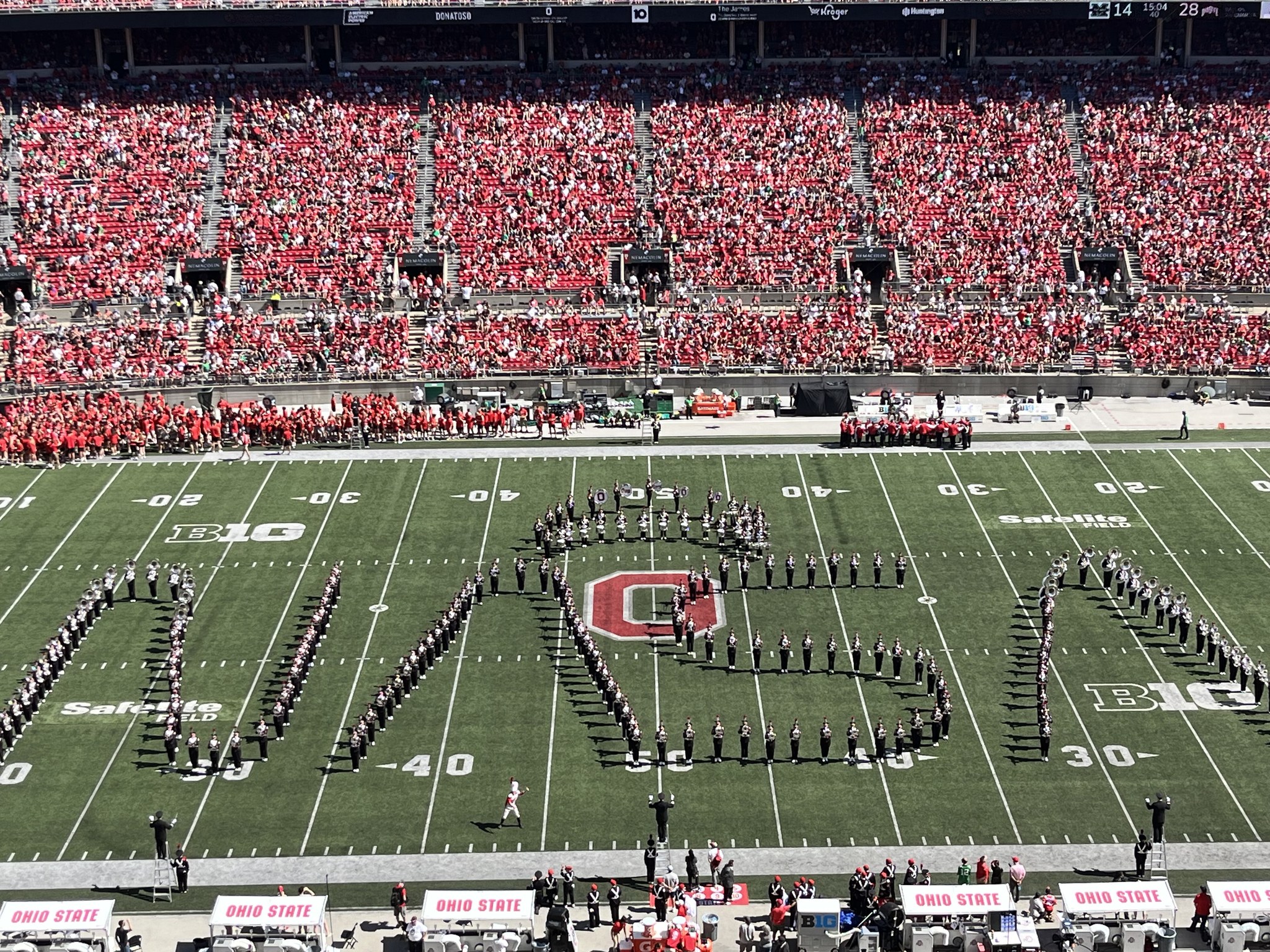 A marching band makes the shape of the NASA worm logo on their football field. Crowds of fans wearing red sit in stands surrounding the field.