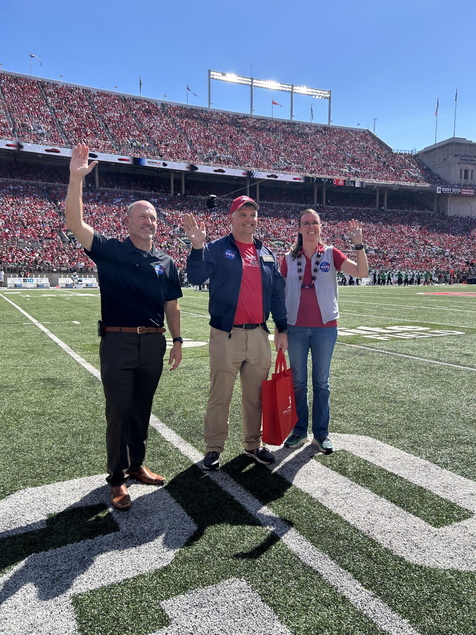 NASA Glenn’s center director and two NASA employees who are Ohio State alums stand on the field and wave. Fans are in bleachers in the background.