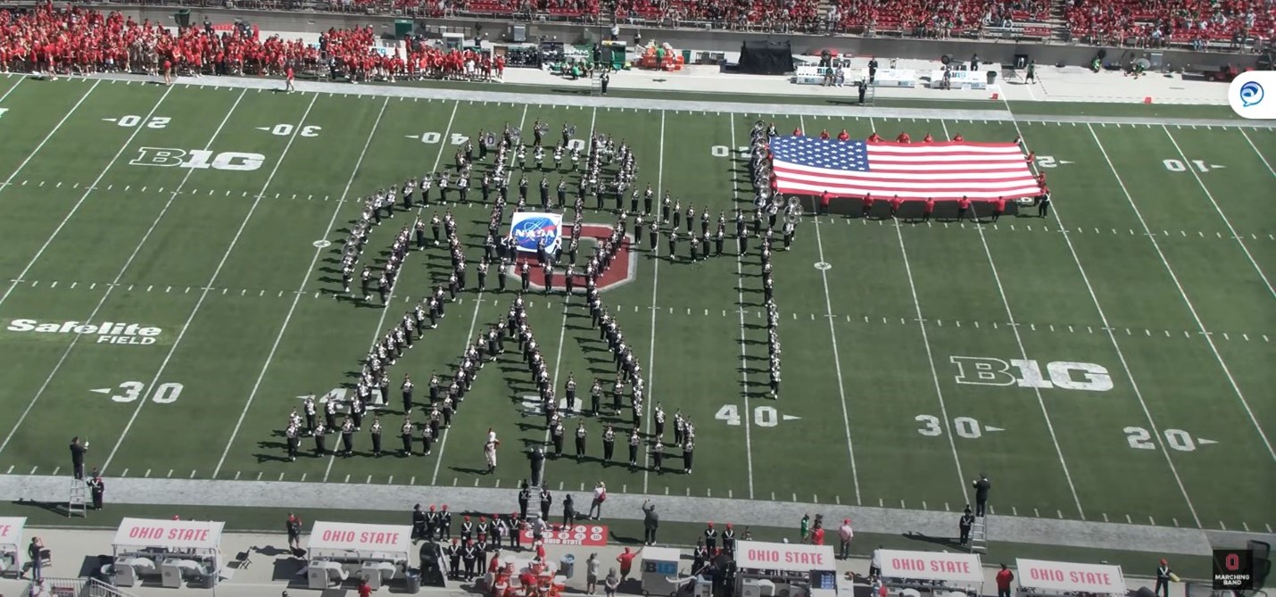 Marching band makes the shape of a large astronaut holding a USA flag on the football field.