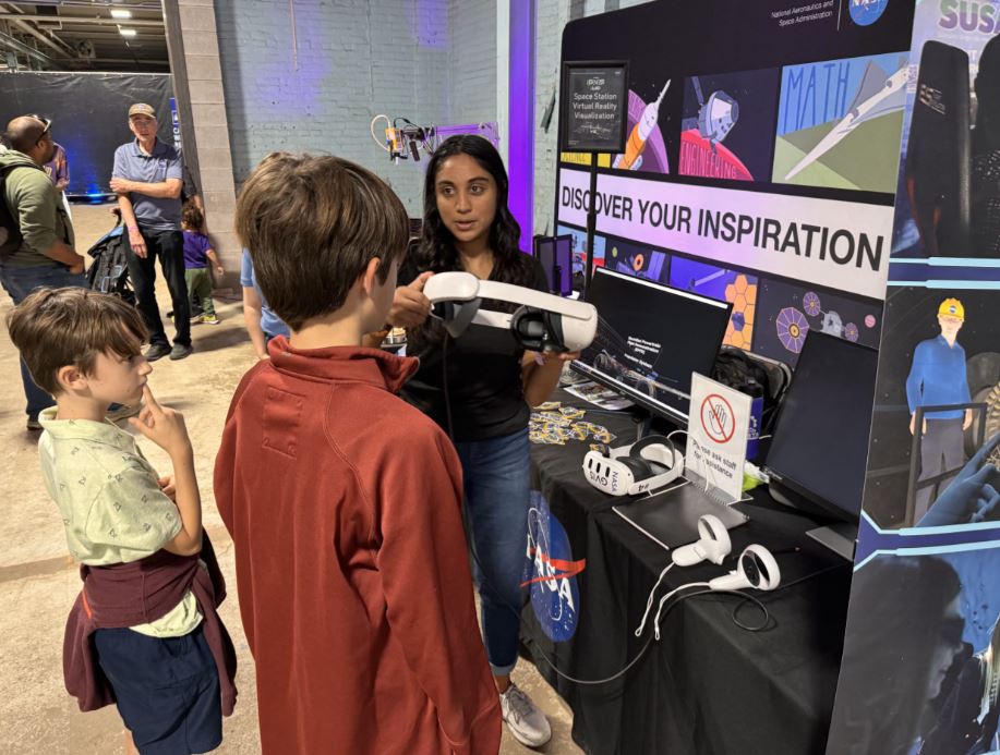 A NASA employee assists a boy with placing an advanced visualization headset on. She stands in front of a display that reads “Discover Your Inspiration.”