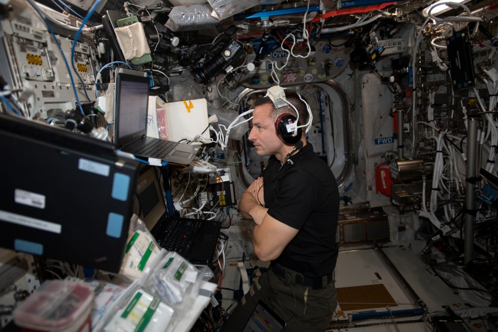 An astronaut wearing headphones works with multiple laptops inside the International Space Station.