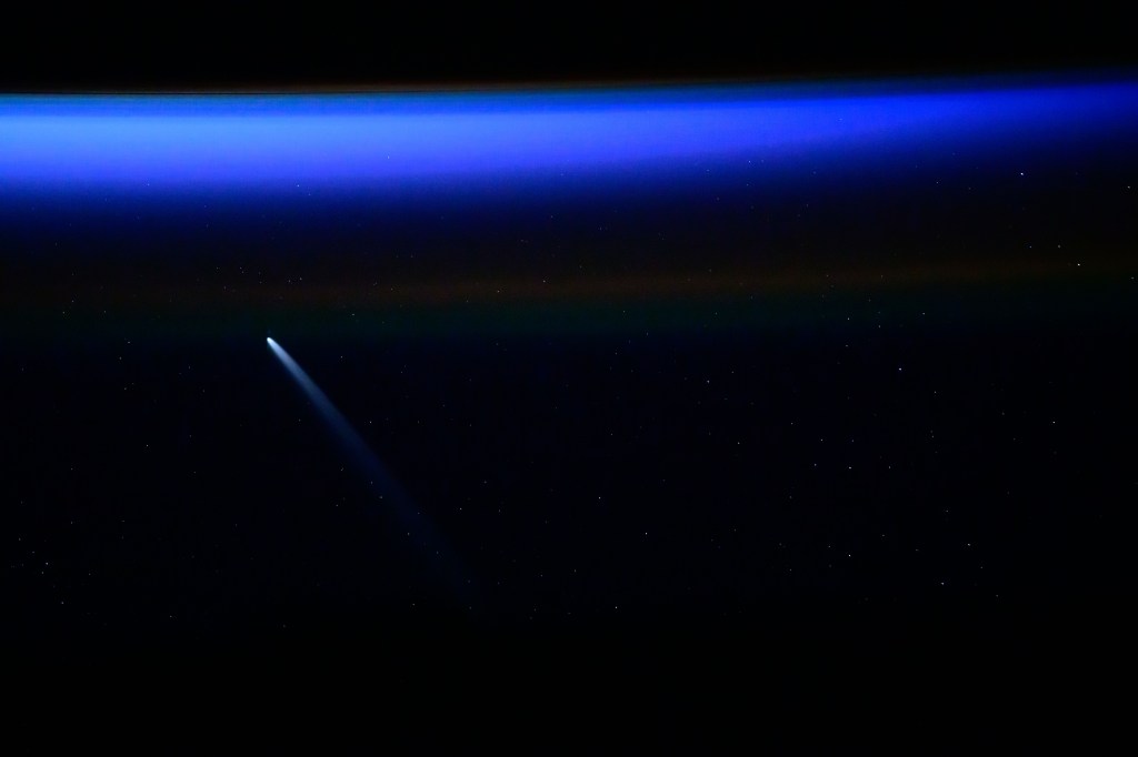 A black background of sky with stars melts into a bright blue horizon color at the top of the image. A comet with a blue trail pictured from the International Space Station as it orbited over 250 miles above the South Atlantic Ocean.
